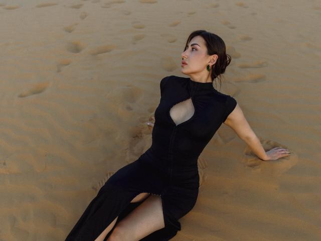 Olivia Cluff, vêtue d'une robe noire avec une découpe, assise dans le sable, les bras derrière elle, les jambes écartées, de petites boucles d'oreilles vertes, des cheveux bruns attachés en chignon, regardant sur le côté..