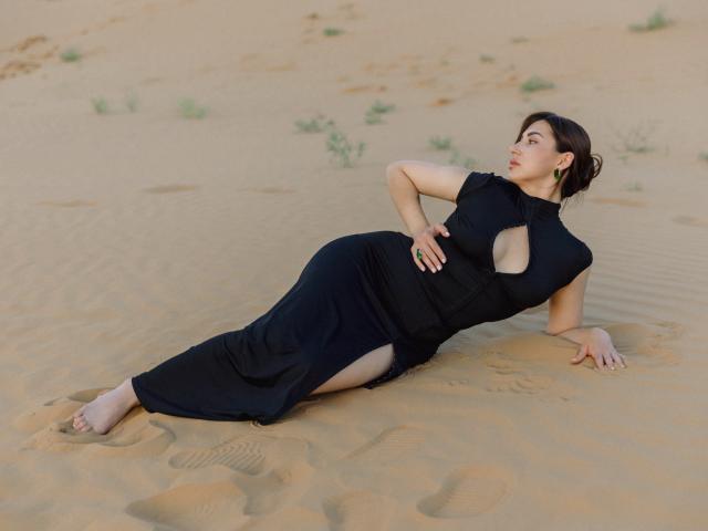 Olivia Cluff allongée sur le sable, vêtue d'une robe noire avec découpes, ses fesses légèrement relevées, cheveux bruns attachés en chignon, boucles d'oreilles vertes, une main sur la hanche, les jambes tendues, regardant sur le côté..