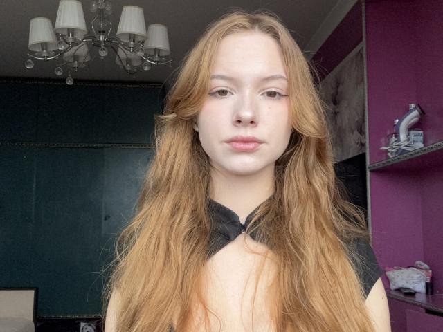 Joette Christianson mit langem, gewellten, blondem Haar, heller Haut und rosafarbenen Lippen, trägt ein schwarzes Oberteil und befindet sich in einem Raum mit einer violetten Wand und einem Kronleuchter. Brüste sind sichtbar..
