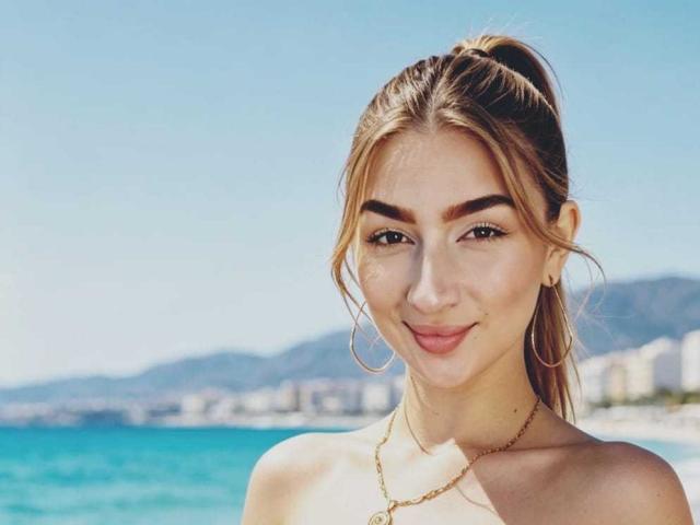AbbyMilers has brown hair in a ponytail, hoop earrings, and a gold necklace, smiling with light makeup, showing her tits, against a clear blue sky and ocean.