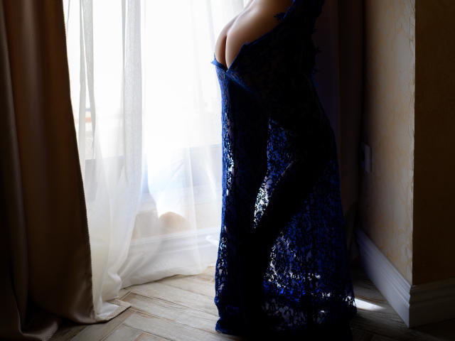 KamiLove, vêtue d'une robe en dentelle bleue, ses seins bien mis en valeur, se tient près d'une fenêtre avec des rideaux blancs, la lumière du soleil soulignant ses courbes. Tissu transparent, sol en bois..