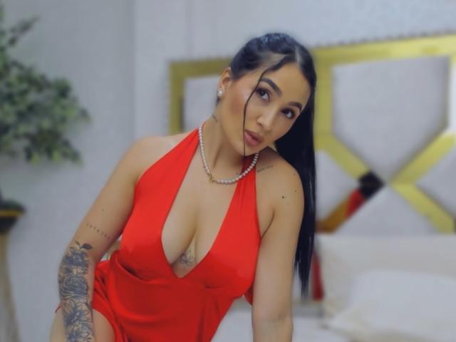 PamelaLiss met lang zwart haar, gekleed in een rode jurk met een lage halslijn, een parelketting, tatoeages op haar arm, en die de camera aankijkt met volgeblazen lippen, waarbij haar grote borsten zichtbaar zijn..