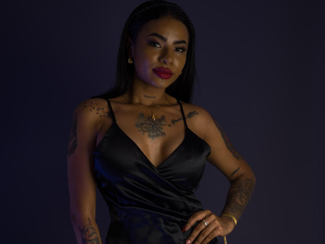 Anneroa, de piel oscura, con cabello negro largo, lápiz labial rojo, tatuajes en el pecho y los brazos, vestida con un vestido de satén negro, un collar de oro y un anillo, de pie con confianza contra un fondo oscuro..