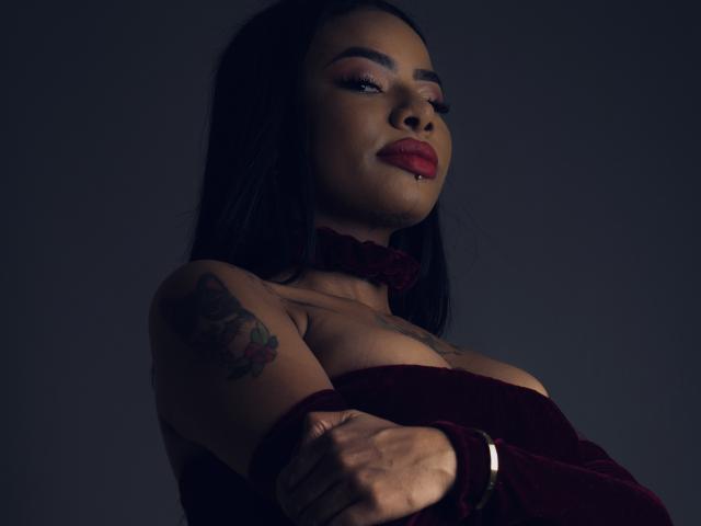 Anneroa, con i capelli scuri, il rossetto rosso e i tatuaggi sul braccio, indossa una collana rossa e un abito dello stesso colore, tiene le braccia incrociate, appare sicura di sé, sensuale e con la pelle scura..