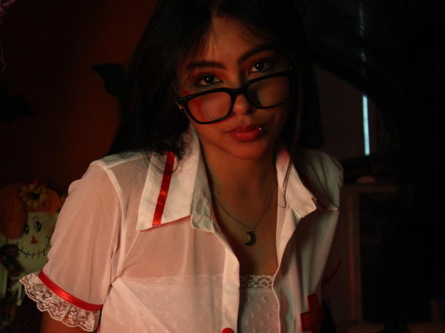 SkyWinter with dark hair, black glasses, and red lipstick, wearing a sheer white nurse outfit with red trim, showing small tits and a lace bra, in dim lighting.