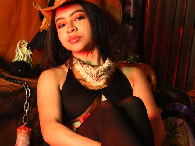 SkyWinter, met donker haar, bruine ogen en rode lippenstift, draagt een cowboyhoed, een zwarte haltertop en een sjaal, zit met de knieën opgetrokken, kijkt naar de camera en heeft een sensuele glimlach..