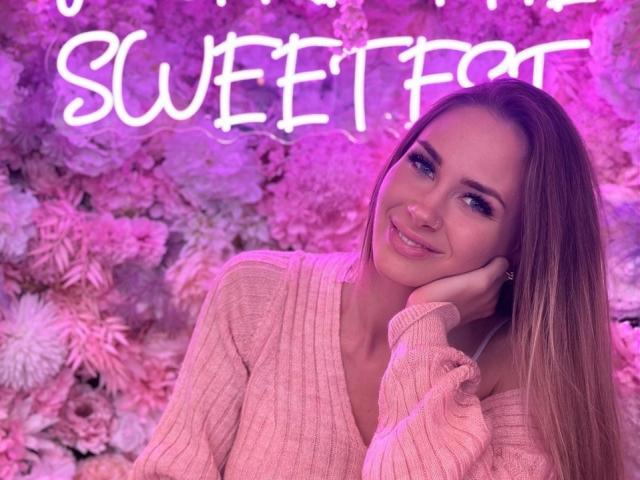 RosalieFleur with long brown hair, pink sweater, smiling, hand on cheek, pink neon "SWEET" sign, pink and purple background, cute, sexy, feminine.