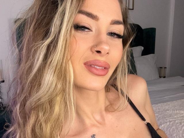 RosalieFleur tiene el cabello rubio, un piercing en la nariz y labios rosados y sensuales. Lleva una blusa negra y tiene un tatuaje pequeño en el pecho. Sus ojos están medio abiertos..
