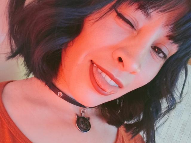 IssisDiosa, avec ses courts cheveux noirs, cligne de l'œil et tire la langue. Elle porte un collier noir avec un pendentif en forme de chat et une chemise rouge. Sa peau est claire..