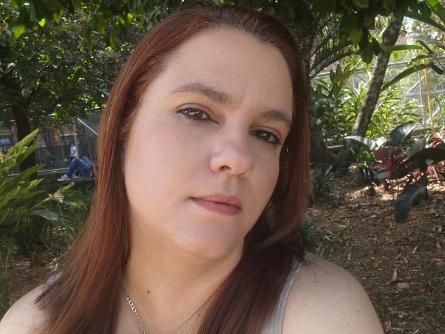 Amelia Wilson tiene la piel clara, ojos marrones y cabello rojo lacio, y lleva una camiseta blanca con un collar de plata. Está mirando ligeramente hacia un lado con una expresión neutra..
