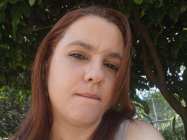 Amelia Wilson tem uma expressão ligeiramente irritada ou cética, pele clara, olhos castanhos e cabelo liso avermelhado, vestindo uma camiseta cinza, sob uma árvore verde..