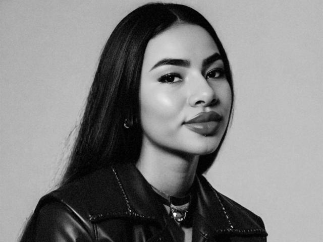 Anneroa tiene el pelo largo y oscuro, labios carnosos, y lleva una chaqueta de cuero con un collar. Está mirando a la cámara con una ligera sonrisa. Foto en blanco y negro..