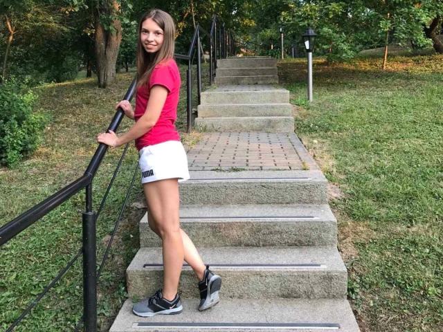 MoonKiss-hot, slim with small tits, wearing a red shirt, white shorts, and black sneakers, stands on stone stairs, holding the black railing, smiling at the camera.