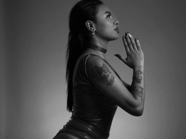 Anneroa, avec sa peau foncée, ses longs cheveux et ses tatouages sur le bras, porte un haut en latex brillant, les mains levées, et regarde sur le côté. En noir et blanc. Les seins, les fesses, le vagin et les tétons ne sont pas visibles..