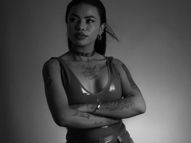 Anneroa mit verschränkten Armen, glänzender Latexoberteil, Choker, Tattoos an Armen und Brust, Ohrringe, blickt zur Seite, Schwarz-Weiß-Foto. Ihre Brüste sind sichtbar. Ihre Vulva ist nicht zu sehen..