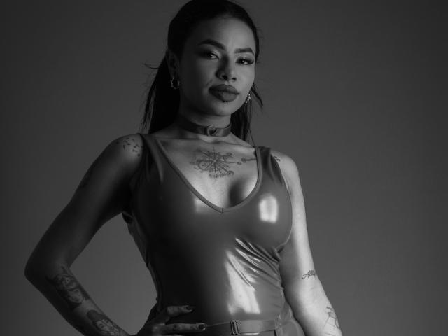 Anneroa met tatoeages, die een glanzend latex tanktop, een choker en oorringen draagt, staat met haar handen op haar heupen, zwart-wit foto, zichtbare tatoeages op de borst en armen, zelfverzekerde uitdrukking..