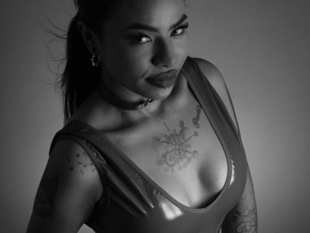 Anneroa, con el cabello oscuro recogido en una coleta, vistiendo una blusa brillante y escotada, mostrando tatuajes en el pecho y los brazos, y un collar, en una foto en blanco y negro..