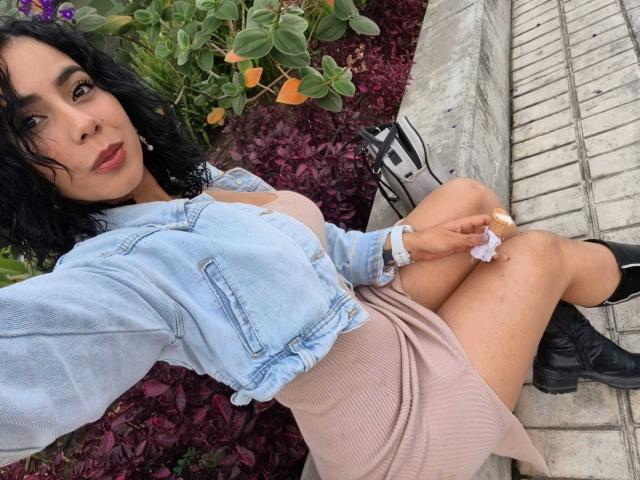 MarianaJackson with dark curly hair, wearing a light blue denim jacket and pink ribbed dress, black boots, and white sneakers, sitting on a brick sidewalk, holding a white scrunchie.