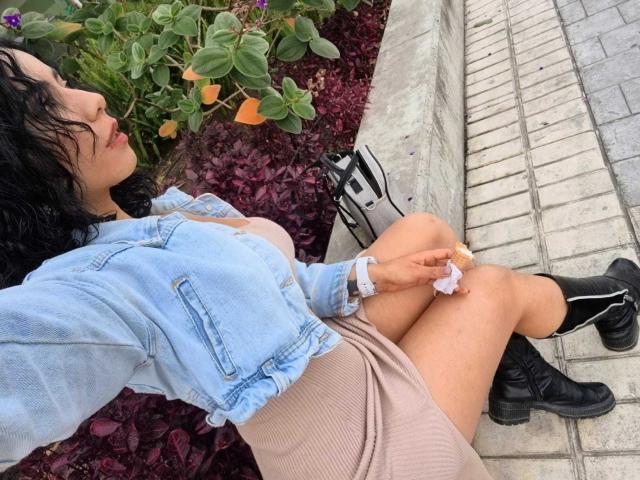 MarianaJackson with wavy black hair, wearing a light blue denim jacket, beige skirt, and black boots, sitting on a tiled sidewalk, eating a small white pastry.