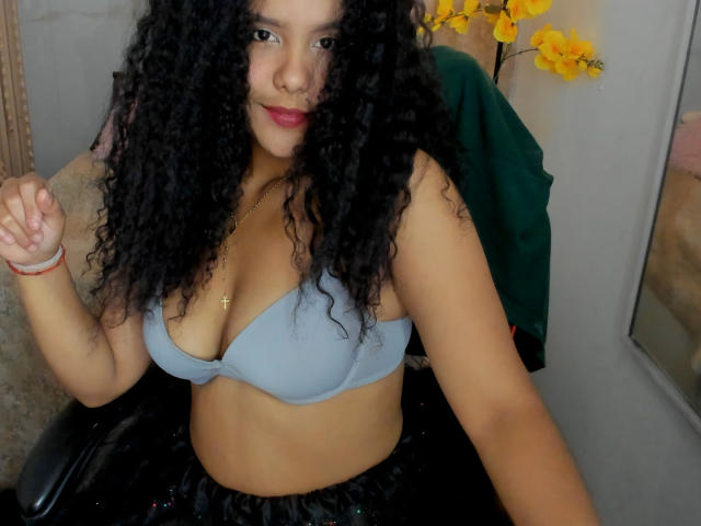 AriaVega with curly black hair, light brown skin, wearing a grey bra, showing her tits, small gold cross necklace, red lipstick, black chair, yellow flowers in background.