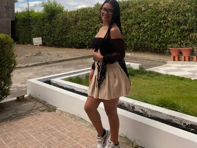 LonxEva, con lunghe trecce nere, indossa una maglietta nera con una spalla scoperta, una gonna beige e scarpe da ginnastica bianche, ed è in piedi su un patio in mattoni, mostrando il suo sedere abbronzato e i suoi piccoli seni..