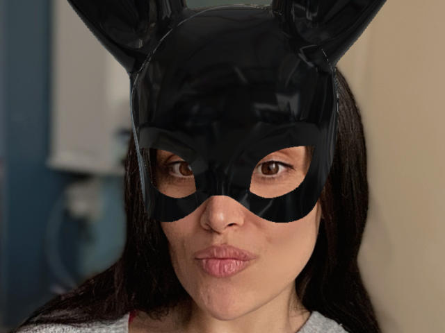 BellaBabeXx porte un masque de lapin noir brillant qui lui recouvre les yeux, ses yeux marrons étant visibles, et elle fait une moue avec ses lèvres roses, affichant une expression légèrement satisfaite..