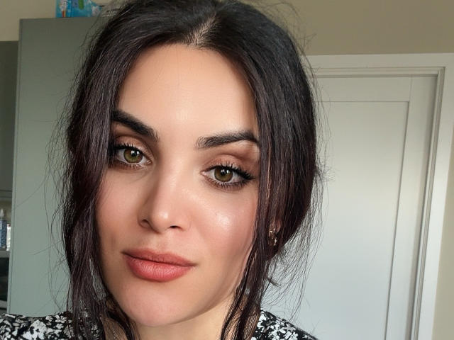 BellaBabeXx a les yeux verts, les cheveux brun foncé, et porte un maquillage discret avec un rouge à lèvres rose. Ses sourcils sont épais, et elle regarde l'appareil photo avec un léger sourire..