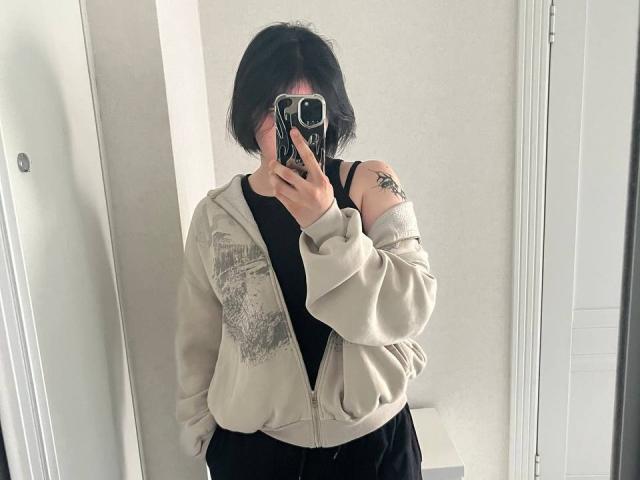 DianaYeess, com cabelo preto curto, vestindo uma jaqueta bege com zíper, uma camiseta preta e calças pretas, tirando uma selfie no espelho com um celular, mostrando uma tatuagem no braço direito..