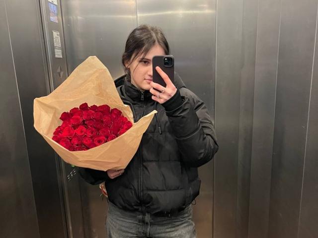 DianaYeess with dark hair in a black puffy jacket, holding a brown paper bouquet of red roses, taking a selfie in a metallic elevator, looking slightly annoyed.