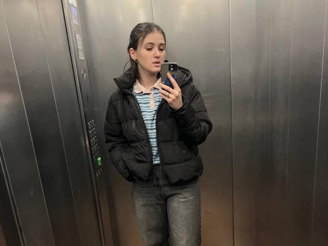 DianaYeess, avec des cheveux bruns attachés en queue de cheval, portant une veste noire matelassée, une chemise rayée et un pantalon gris, prend un selfie dans un ascenseur en métal, les mains dans les poches..