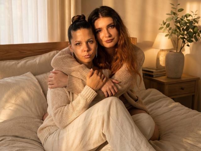 WillodeanLimes mit braunen Haaren, die zu einem Dutt gebunden sind, trägt einen beigen Pullover und sitzt auf einem Bett, die Arme um eine dunkelhaarige WillodeanLimes gelegt, die einen ähnlichen Pullover trägt. Beide schauen in die Kamera..