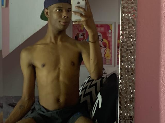 AndyCris, torse nu, mince, peau foncée, fesses petites, seins petits, portant un short gris, une casquette bleue, tenant un téléphone, autoportrait devant un miroir, souriant, tatouage sur le bras gauche, oreiller noir et blanc..