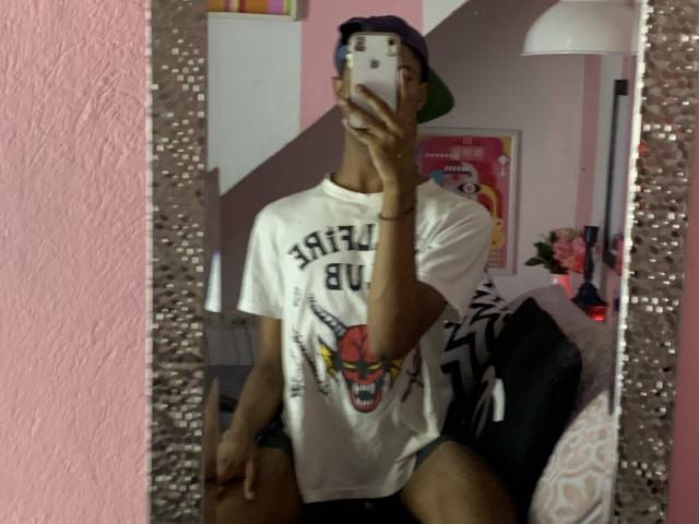 AndyCris portant un t-shirt blanc avec un motif de visage de tigre, un short noir et une casquette à l'envers, prenant un selfie devant un miroir, montrant une silhouette mince et une peau foncée..