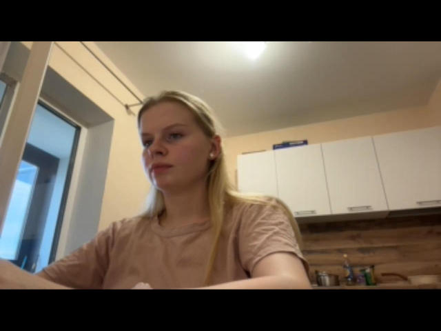 RendaKu hat blondes Haar, helle Haut und trägt ein pinkes Hemd. Sie blickt leicht nach links und hat einen neutralen Gesichtsausdruck, und befindet sich in einer schlecht beleuchteten Küche..