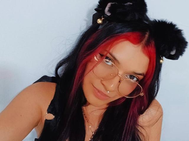 AriaRosel with black and red hair, wearing cat ears and gold glasses, has a nose piercing and is smiling at the camera, showing her tits in a black top.