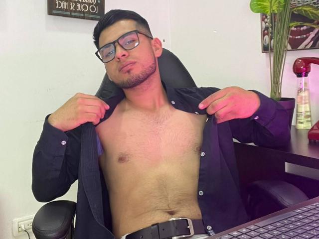 ThiagoVegga, de óculos, com camisa preta aberta, mostrando o peito peludo e os músculos abdominais, rosto levemente emburrado, mão direita na gola da camisa, mão esquerda no teclado. Ambiente de escritório, cadeira preta..