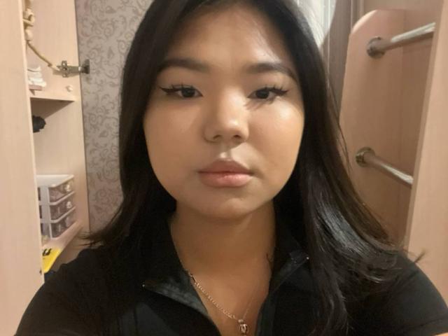 EllaBadgero has a cute, serious face with long black hair, wearing a black shirt and a gold necklace, with thick eyelashes and a neutral expression.