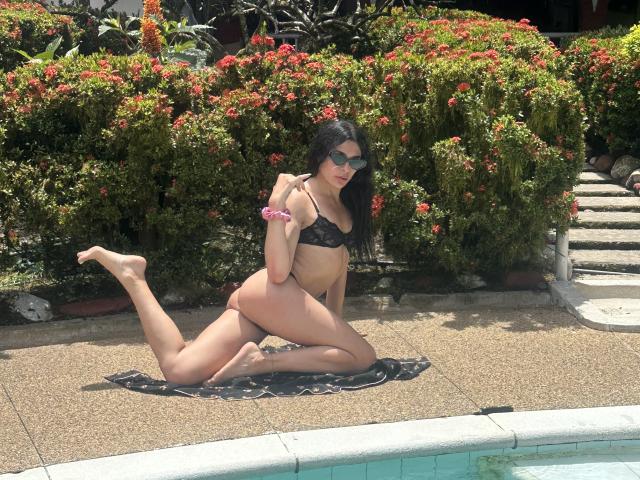 ArielNitzche, kneeling by the pool, wears a black lace bikini, showing her ass, with her legs spread and one knee on the ground, sporting sunglasses and a pink bracelet.