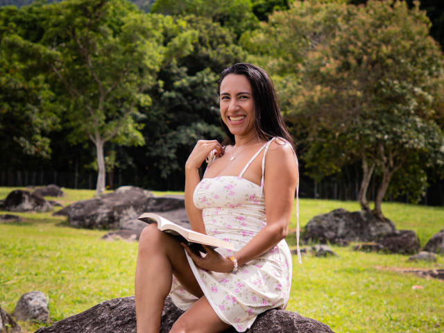 JadeFoster, con i capelli scuri e un sorriso carino, è seduta su una roccia, indossa un vestito floreale, tiene in mano un libro, mostrando il seno e i glutei, in un parco soleggiato..