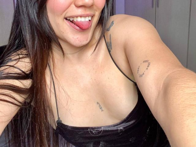 SophiaMarx with long dark hair, tongue out, smiling, showing tattoos on her arm and chest, wearing a black spaghetti strap top, exposing her tits.