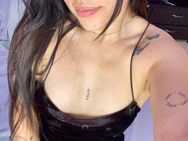 SophiaMarx with long black hair, wearing a black spaghetti strap top, showing her tits and tattoos on her chest and arm, slightly smiling with pink lips.