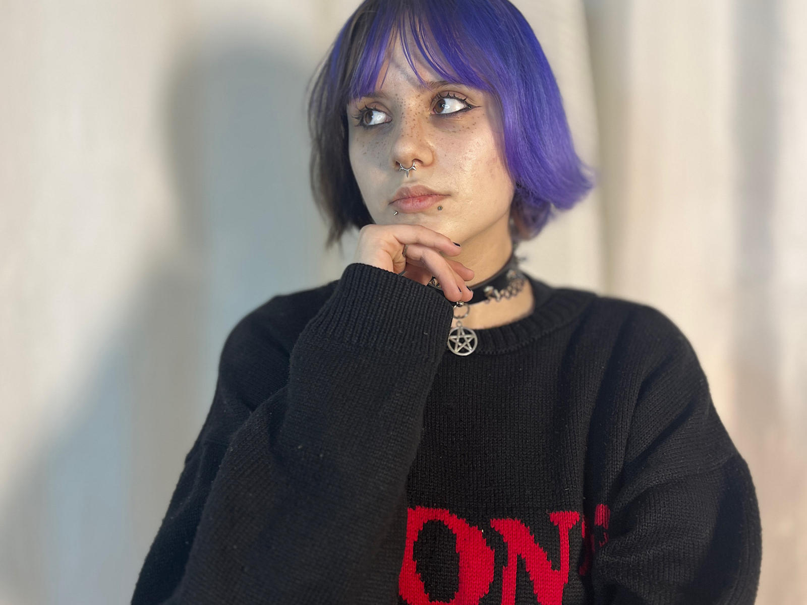 YasukoCella, con cabello morado, aro en la nariz negro, pecas, collar y un suéter negro con letras rojas, pensativa, con la mano en la barbilla..