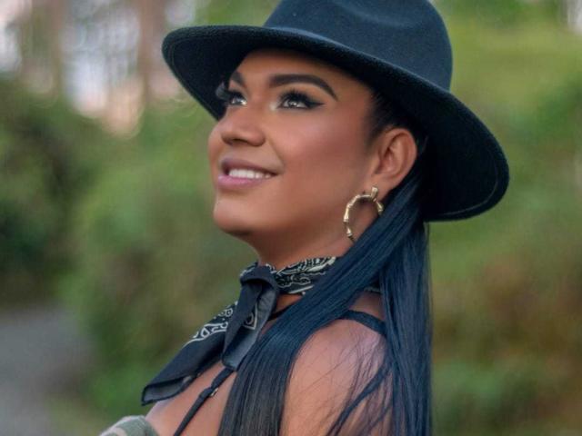 AmberBrownn ha la pelle scura, lunghi capelli neri e indossa un cappello nero con un orecchino dorato a cerchio, sorridendo con il trucco e una bandana nera intorno al collo..