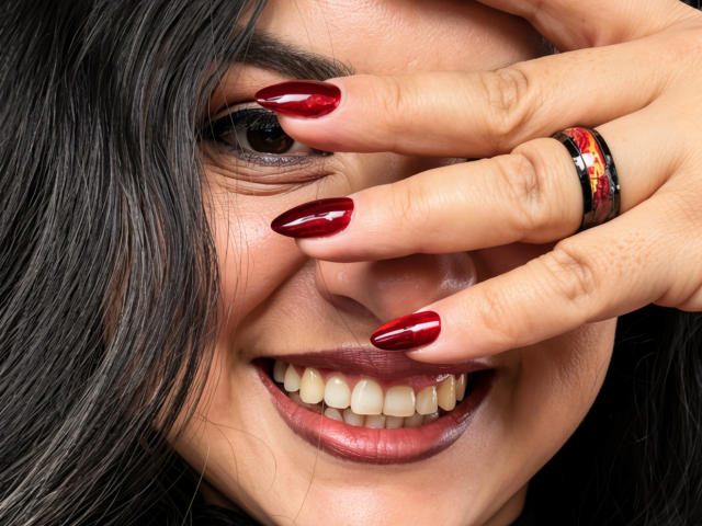 Il viso di Charlotte Devereaux, con i capelli scuri, le unghie rosse e lucide, un anello nero con un motivo rosso e la bocca leggermente aperta che mostra i denti bianchi. Sta coprendo un occhio con la mano..