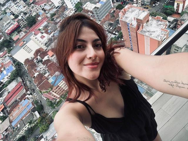 SpencerLe, con cabello castaño y una blusa negra, mostrando un tatuaje en su brazo derecho, sonriendo a la cámara, con un horizonte urbano de fondo, tomándose un selfie desde un lugar elevado..