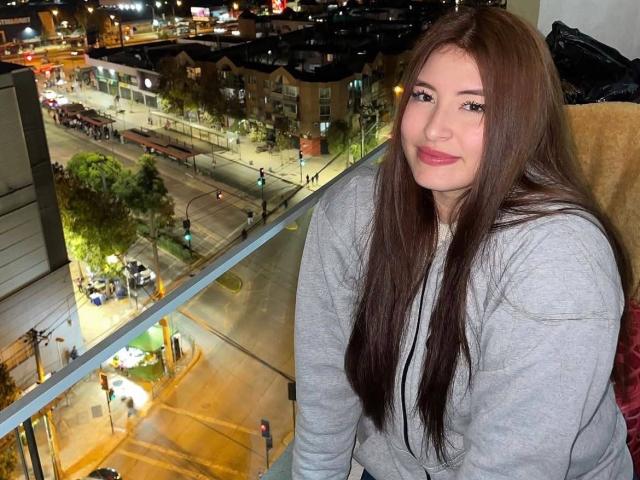 CassiBell, avec de longs cheveux bruns, un sweat à capuche gris et un sourire, assise près d'une fenêtre la nuit, avec les lumières de la ville et la rue visibles en contrebas, a l'air mignonne et détendue..