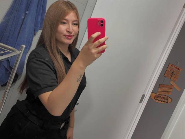 CassiBell, con lunghi capelli castani, una maglietta nera e un telefono rosa, sta scattando un selfie allo specchio, mostrando un tatuaggio sull'avambraccio destro, sorridendo con rossetto rosa, in un bagno..