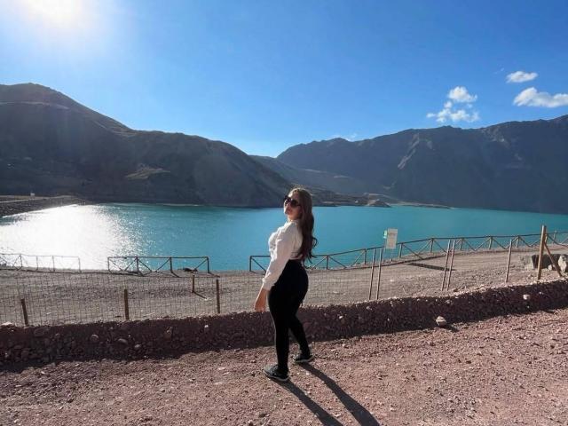 CassiBell, de espaldas a la cámara, lleva una camiseta blanca, pantalones negros y zapatos negros, con el pelo largo y castaño, y está mirando un lago azul brillante y montañas..