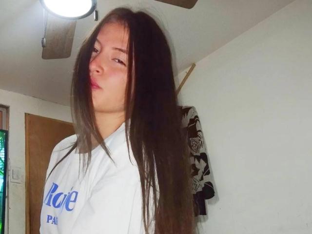 CassiBell, con el pelo largo y castaño, labios fruncidos, vestida con una camisa blanca, está de pie en una habitación con un ventilador de techo y una puerta de madera. Sus pechos son ligeramente visibles debajo de la camisa..