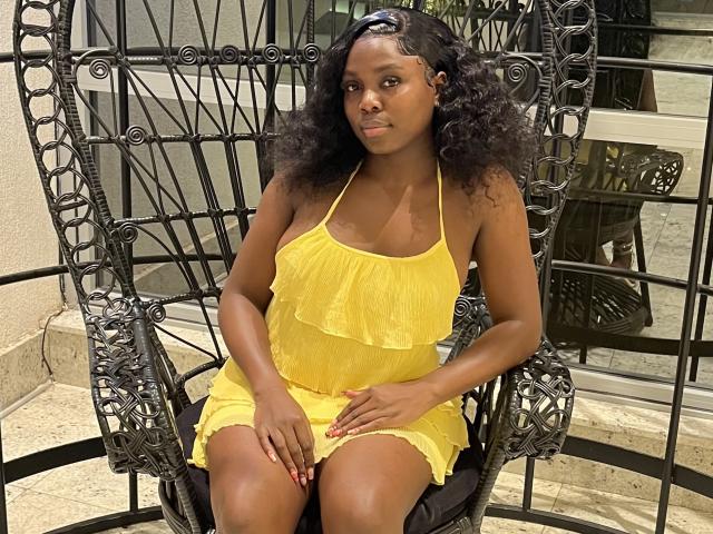 SmoothChocolate mit dunkler Haut, lockigem schwarzem Haar, trägt ein gelbes Top mit Trägern, sitzt in einem verzierten schwarzen Stuhl, die Hände auf ihren Oberschenkeln, blickt leicht zur Seite, rote Fingernägel..